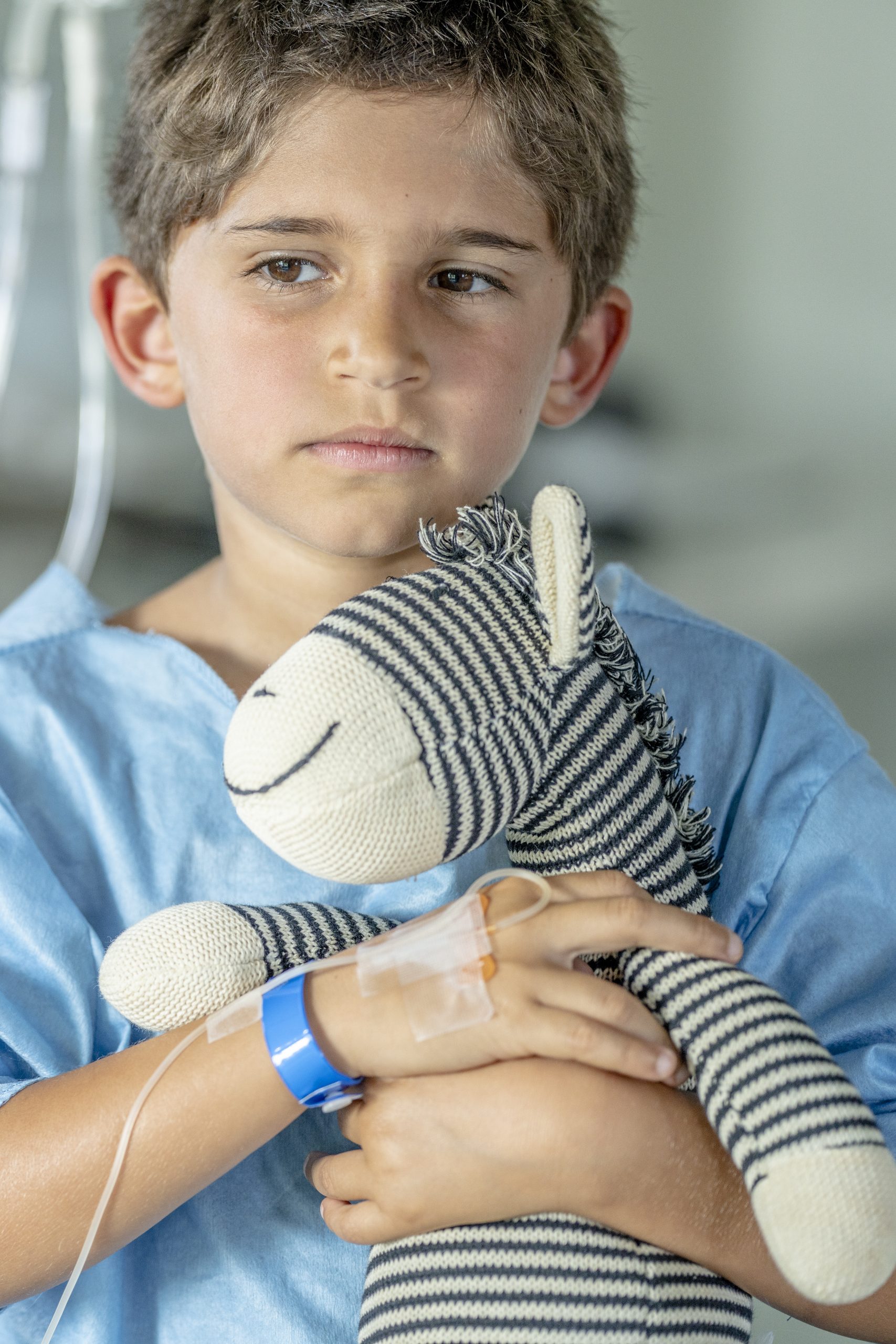 Close up of a six year old boy sitting in the hospital with an IV drip holding his favorite stuffed animal and looking concerned