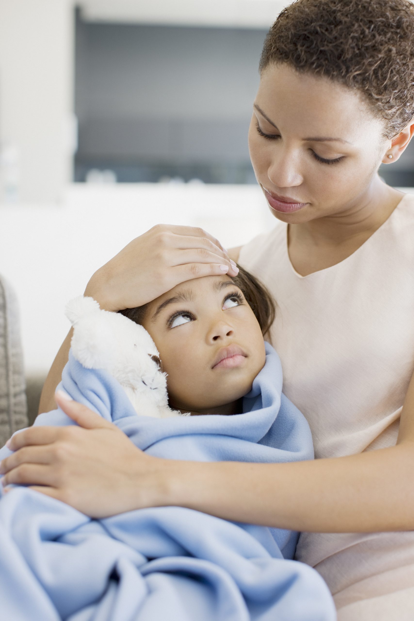 Woman touching the forehead of and looking down at sick daughter in her arms. Her daughter is wrapped in a blanket holding a stuffed animal and looks up at her with big eyes