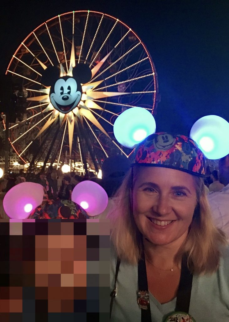 Photo of Dr. Armer with light up ears in front of Pixar Pal-A-Round, a swinging ferris wheel, in California Adventure with her child whose identity is protected with pixelation