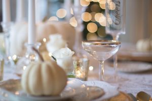 Table decorated for a special occasion with gold rimmed wine glass, a tall flute that says "Cheers" in gold, a cream colored squash and a plate setting, a white rose flower in a glass and a white candle in the background