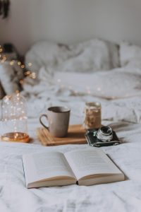 Cozy atmosphere of a bed with a book, a camera leaning against a tray with a coffee cup and a candle, and fairy lights in a jar and in the background