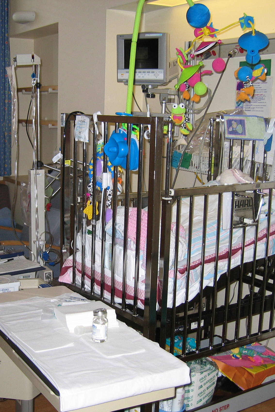 A hospital room with a bay window seat and a metal crib for a baby which has baby blankets in it and a mobile attached with a rolling table next to it that has medicine container and a checklist with a pen