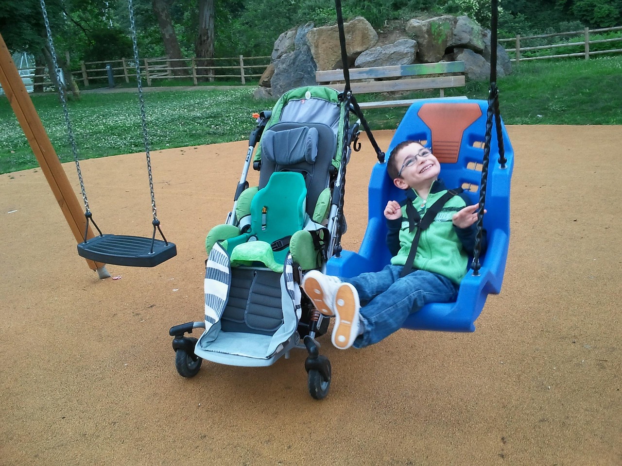 Young boy with blue glasses stretching his feet out while buckled in and swinging in a large blue plastic adaptive swing. His supportive adaptive stroller with modifications to support his head and torso is parked right next to the swing.