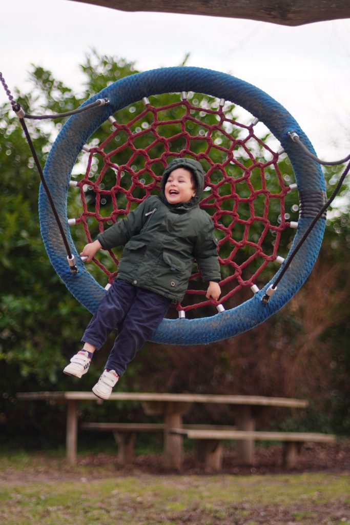 Young boy in a puffy hooded coat smiling and holding on tight while laying flat and swinging on a spider web tree swing featuring a durable, blue-covered steel frame and a vibrant red rope web